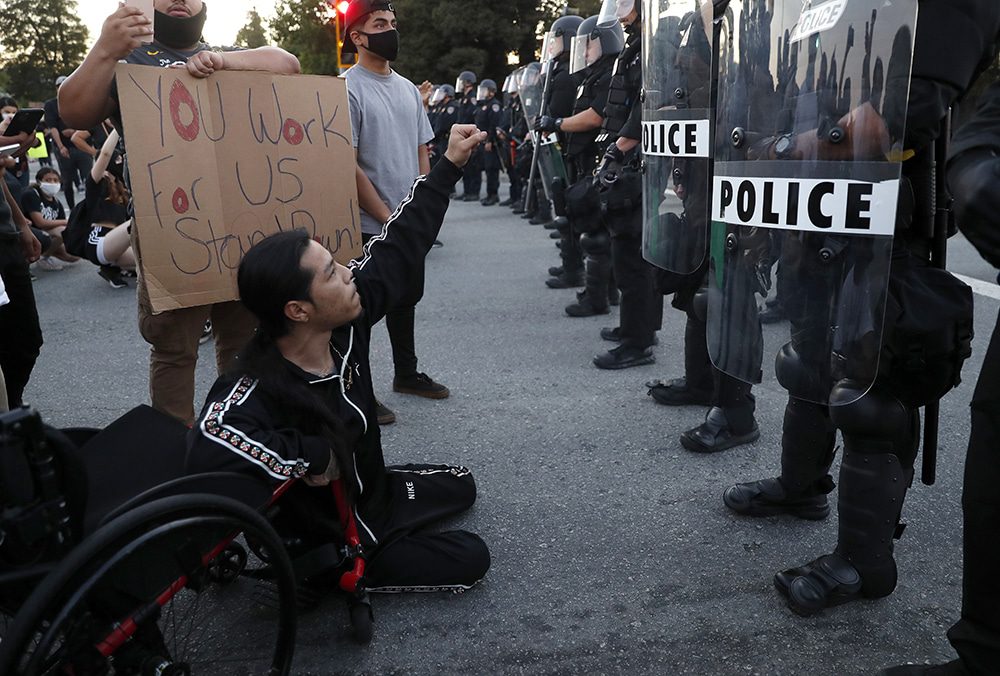 A man sitting on the ground next to his wheelchair holds up his fist in protest against a line of police officers wearing riot gear. A man beside him holds a sign that says "you work for us, stand down!" Digital Accessibility